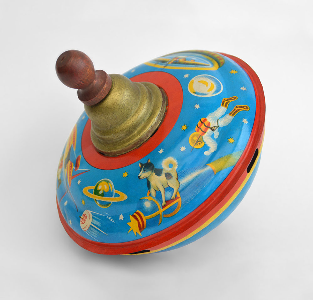 Made by S. I. Toys, this spinning top depicts Laika standing on a version of Sputnik 2 surrounded by a space-themed frieze (1958)